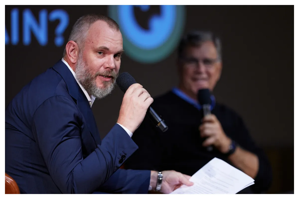A man with a thoughtful expression speaking at a conference, the room has a modern, clean aesthetic with cool blues and muted grays.