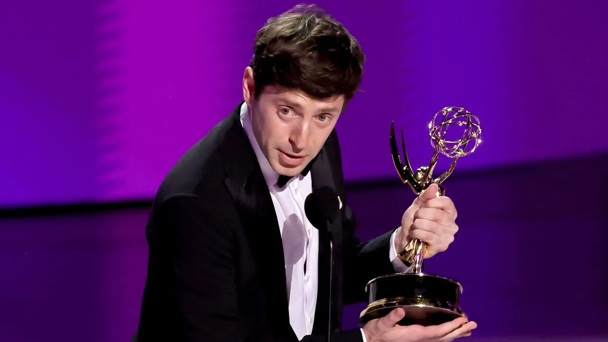 Comedian Alex Edelman standing on a stage with a microphone, looking thoughtful.